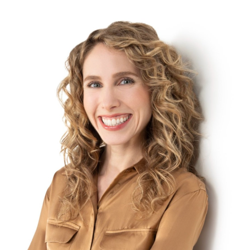 A person with long curly hair and a warm smile, wearing a brown blouse, stands against a white background.